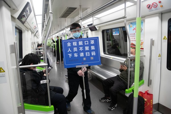 武汉市地铁部分线路星期六恢复运营，但站台候车及车厢内乘客较少。一名地铁执勤员手拿着“全程请带上口罩”、“勿群聚”等牌子，而乘客必须扫码进出车站。（欧新社）